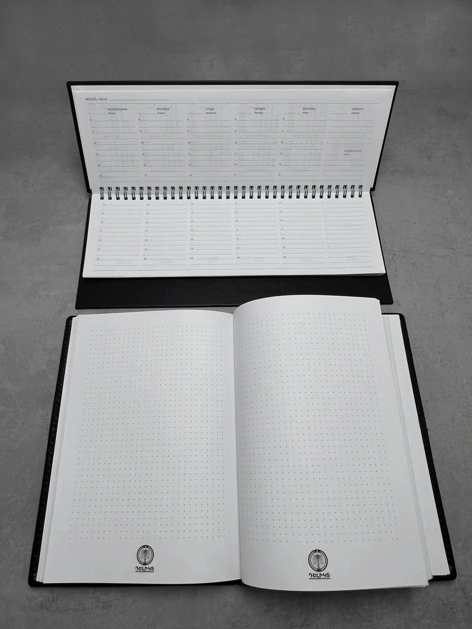 An open notebook with dotted pages and a spiral-bound desk planner. The photograph shows the printed pages, binding construction and branded details.