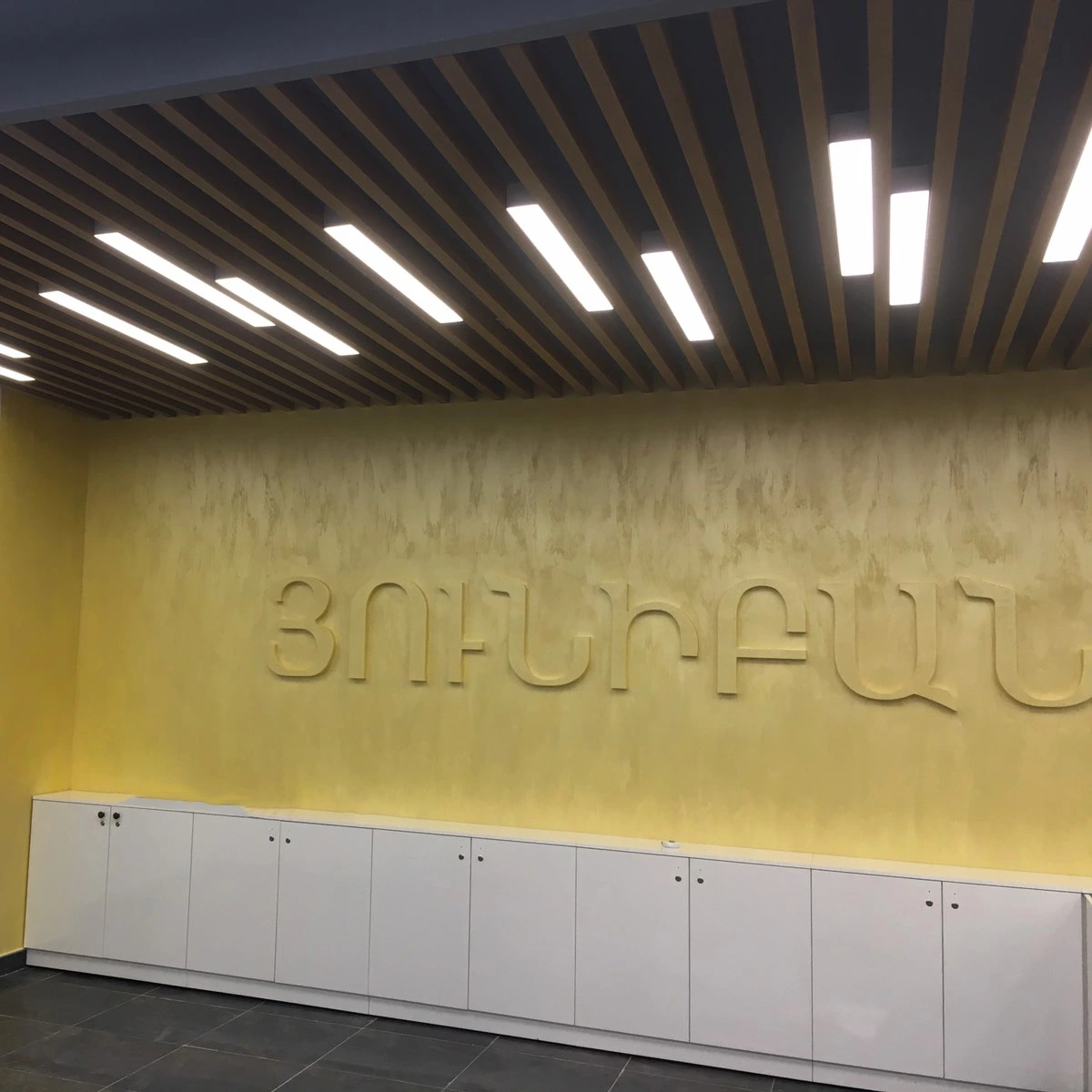 Unibank interior branding with a textured yellow wall, dimensional Armenian lettering and a ceiling with wooden elements. A photograph of the completed project.